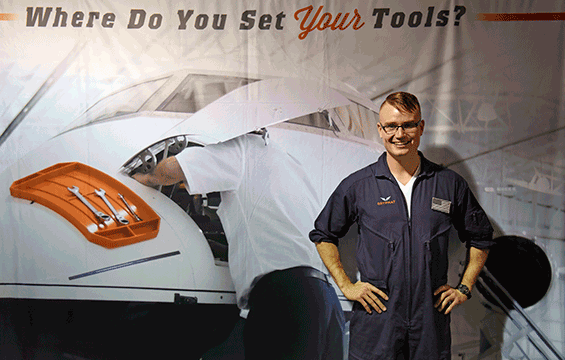 Tom Burden, an F-16 weapons mechanic assigned to the 180th Fighter Wing, stands in front of a banner for his invention.