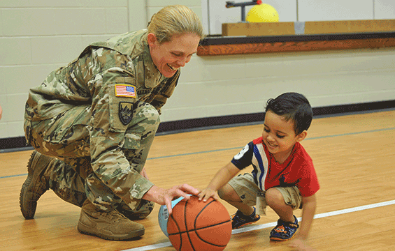 Solider kneels on floor next to child playing with basketball.