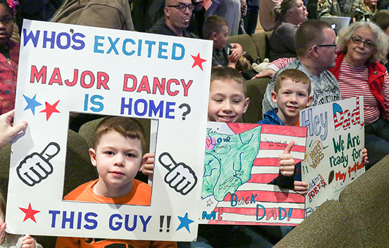 Boys hold signs while they wait for their Dads to arrive from deployment. One sign reads "Who's excited Majoy Dancy id home? This Guy (photo of boy)!!