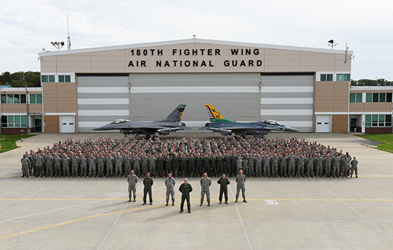 Group shot outside of 180th Airmen with F-16s in back.
