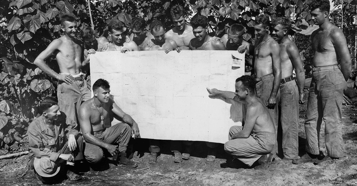 Black and white photo of shirtless Soldiers looking at large chart.