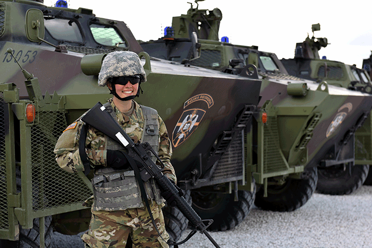 Female Solider in gear standing in front of military vehicles.
