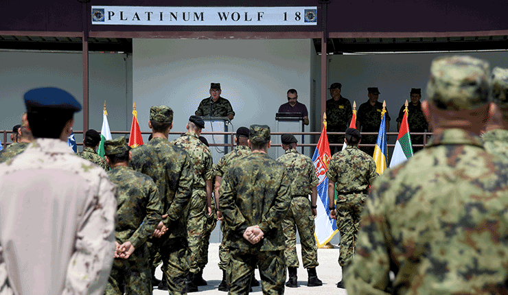 Soldiers facing raised platform stand watching presenters.