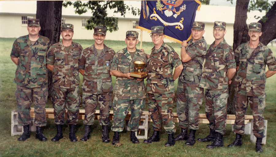 Group of Soldiers pose with award outside with organizational flag displayed.