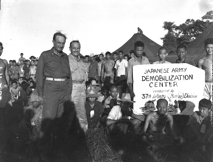 Maj. Gen. Robert S. Beightler poses with Japanese prisoners near sign at demobilization center.