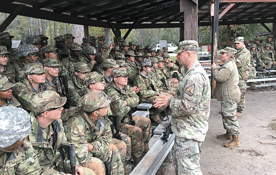 recruited Soldiers sit on bleachers under awning listening to recruiters talk.