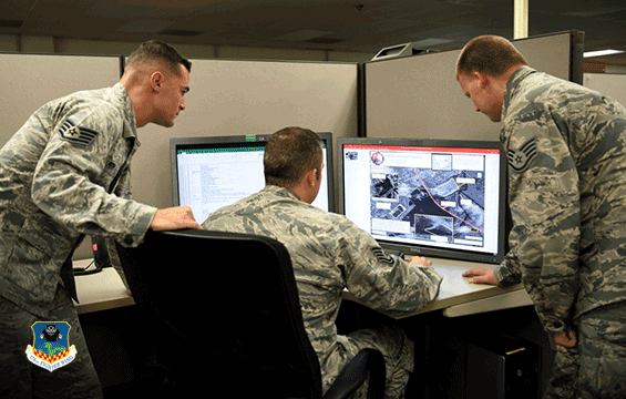 Three airmen galer around desk to look at computer screen.