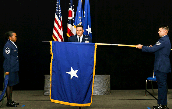 Airmen stand to left and right (holding one-star flag) of podium with BG Camp at podium in center.
