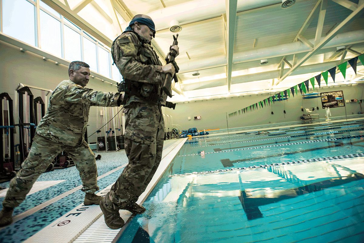 Airman pushes a blindfolded airman holding weapon into a swimming pool. 