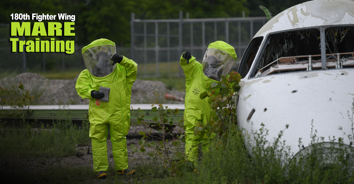 Airmen in chemical suits stand next to a simulated wrecked airplane
