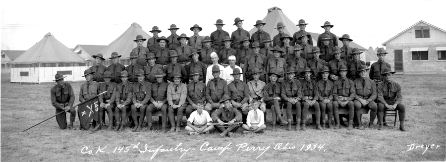 Black and white photo of group sitting in rows outside camp.
