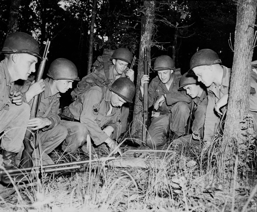 Black and whote photo of soldiers kneeling in woods as they go over plans.