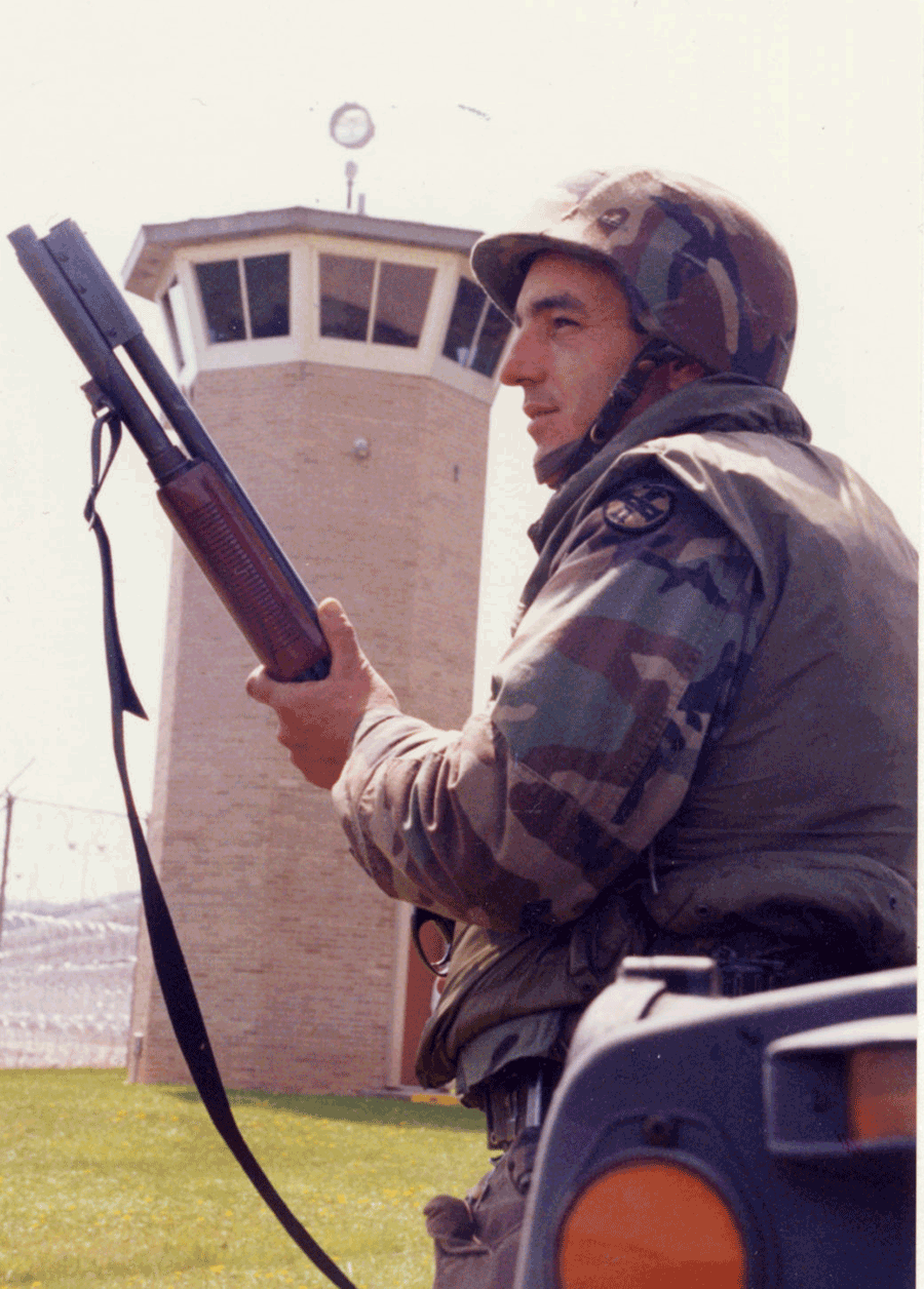 Soldier stands profile with weapon with guard tower in background.