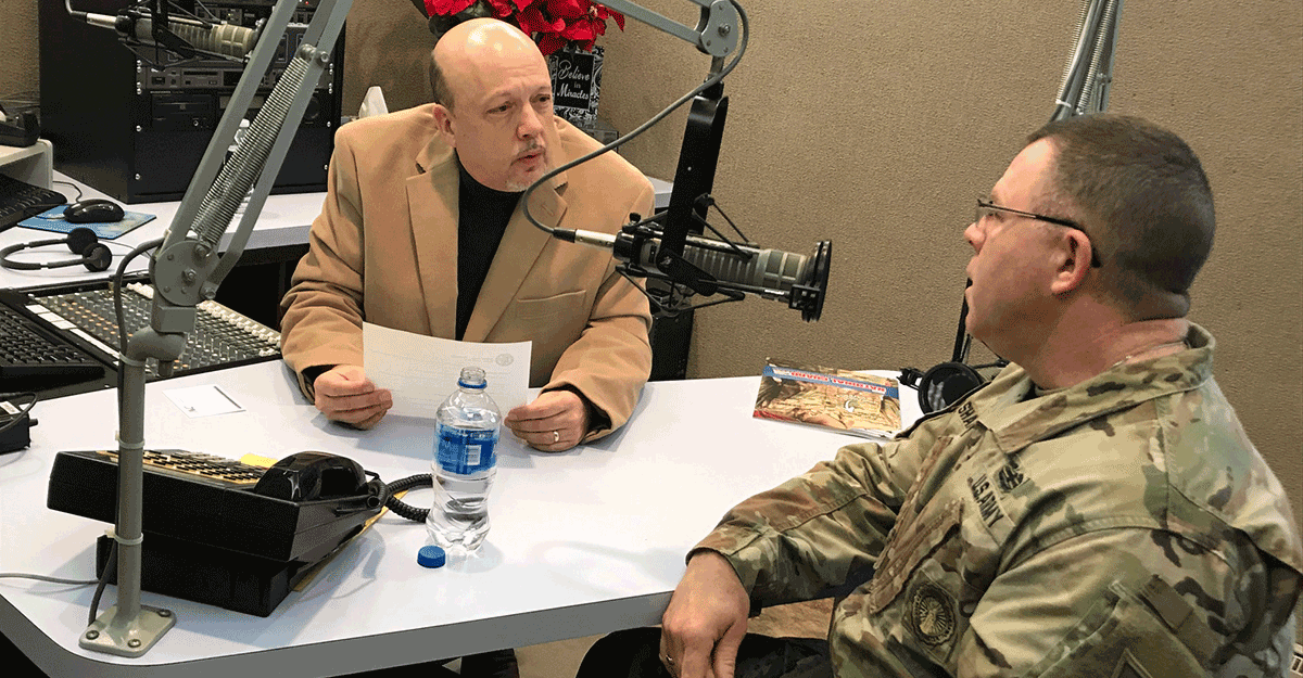 News anchor and Col. Daniel Shank sit at microphones at radio station. 
