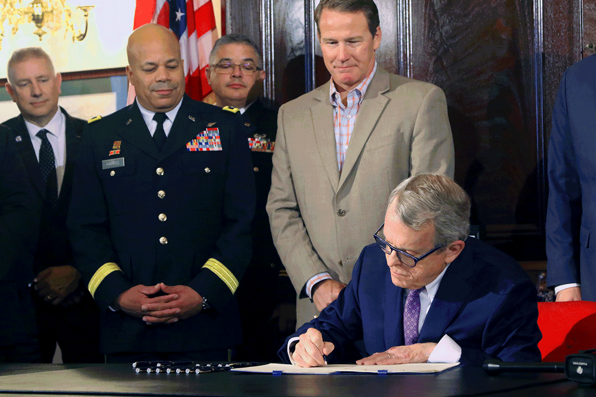Governor Mike DeWine signing legislation with The Adjutant General and others standing behind desk.
