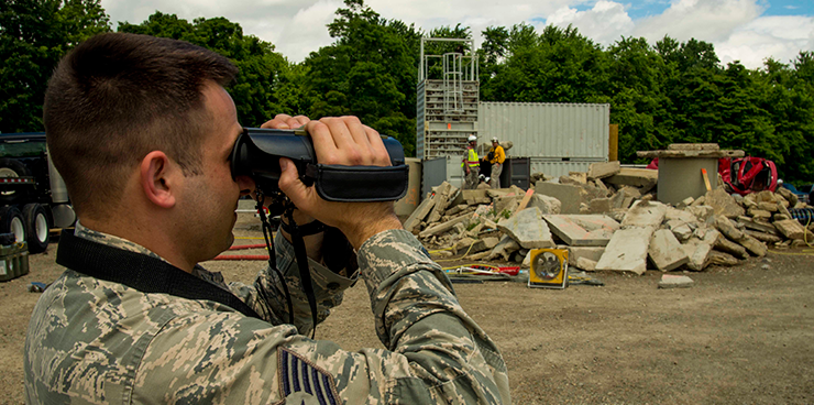 Airman looks through binoculars at concrete rubble site.
