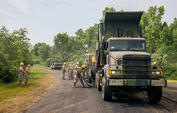 Camo truck dumping asphalt while guard members work it on road.