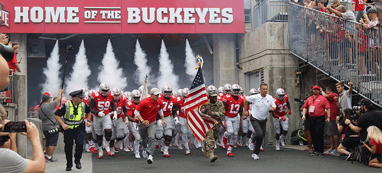 Ohio State Buckeyes run out of tunnel onto field with coach Urban Meyer, led by Soldier running with American flag.