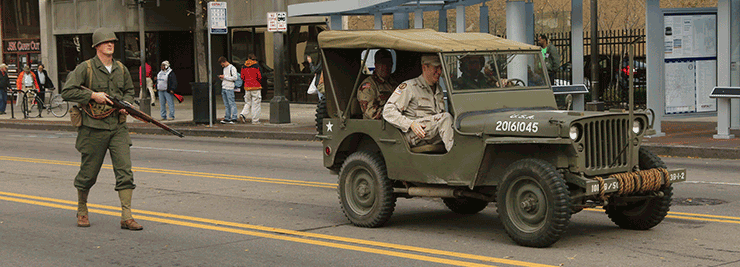 Sgt History (aka Sgt. 1st Class Joshua Mann) marches in parade alongside jeep.