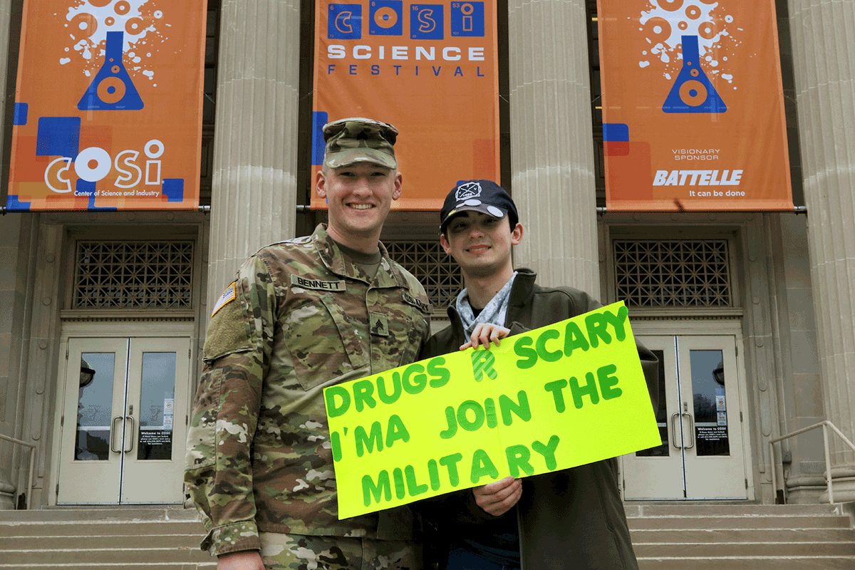 Soldier stands with student holiding sign that reads 'DRUGS R SCARY I'MA JOIN THE MILITARY' prior to march on steps of COSI. 