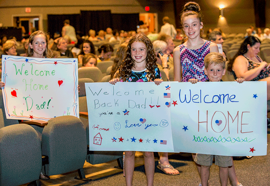 Kids stand in church isle holding up signs that say 'WELCOME HOME DAD'. 