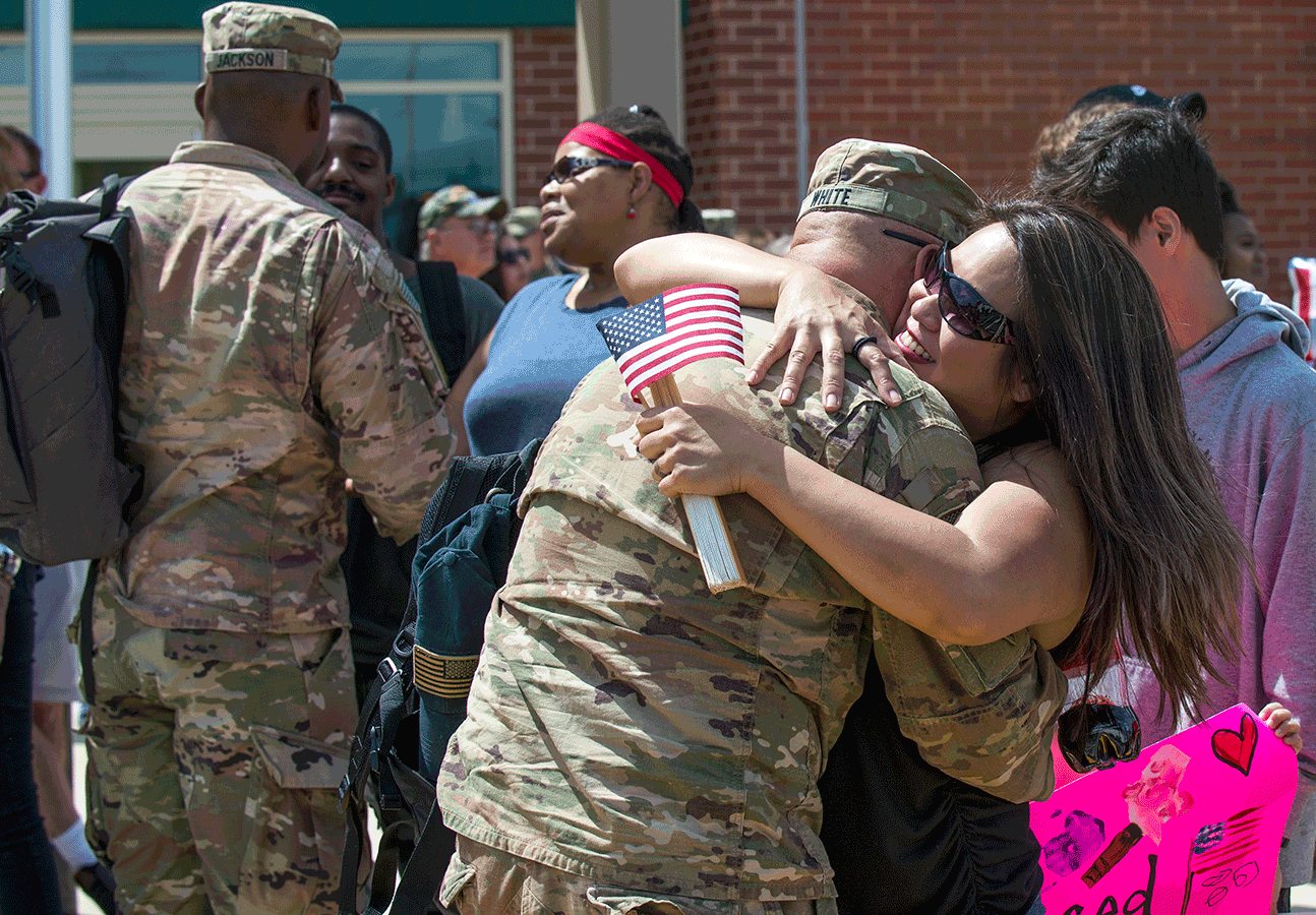 Uniform Soldier hugs wife while other uniformed Soldiers greet family and freinds.