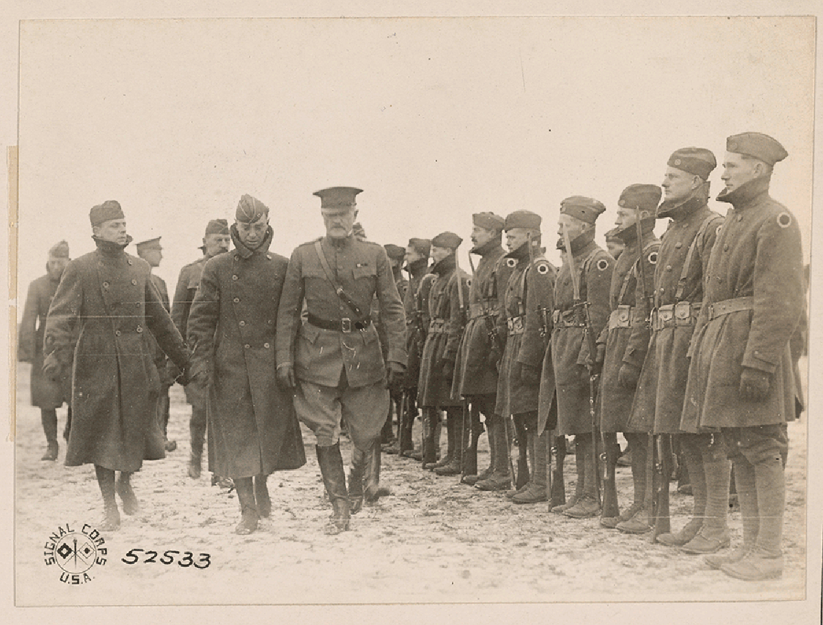 cepia-tone photo of General walking through snow with other officials in front of Soliders standing at attention