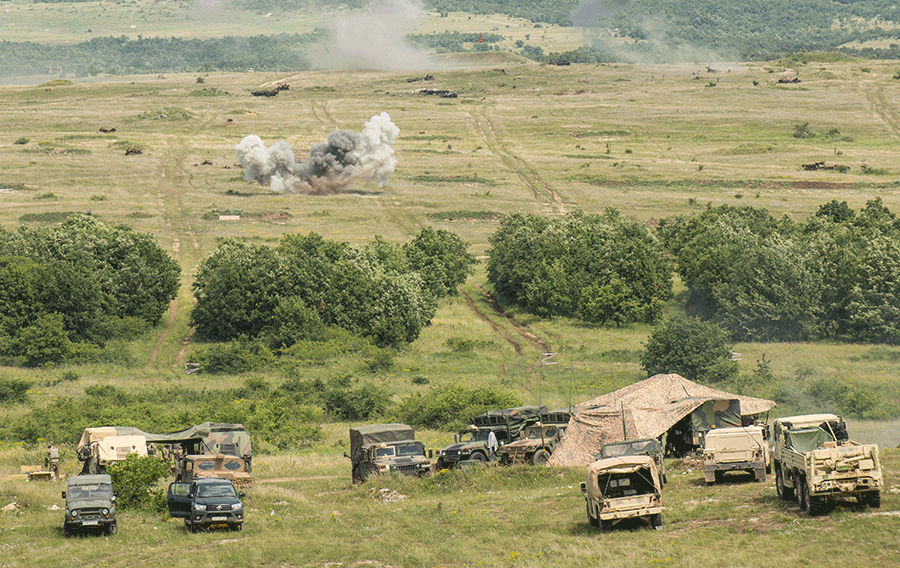 Fields where tents and army vehicles are stationed with explosions in the distance.