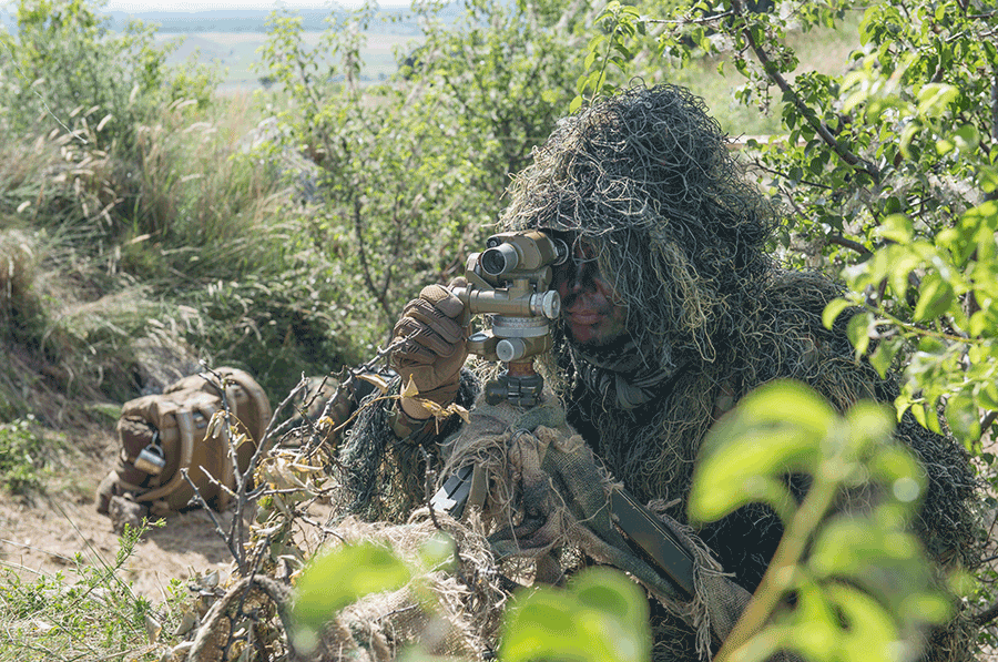 Soldier in ghillie suit on ground looks through scope.