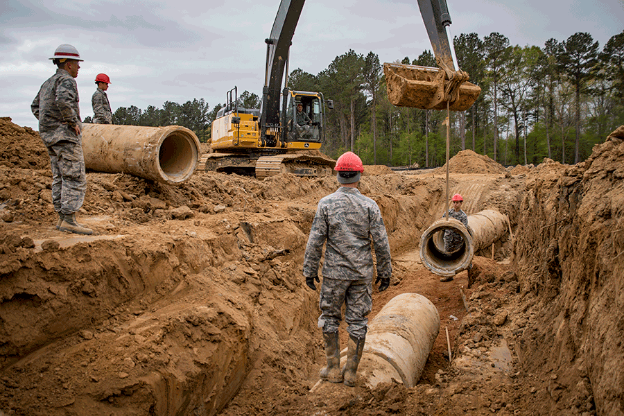 Airmen in ditch, awaiting next section of concrete sewer pipe being lowered by crane.