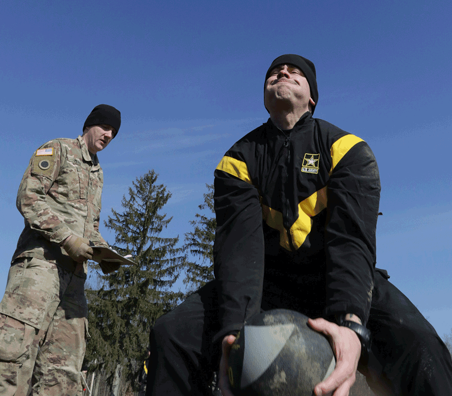 Soldier squatting as he prepares to throw the medicine ball while uniformed Soldier watches.