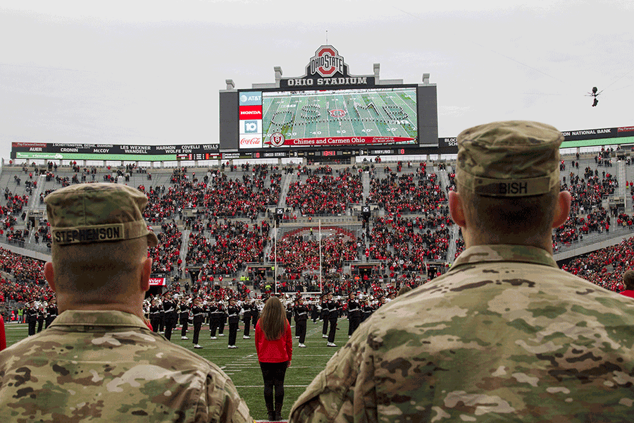 Soldiers and Airmen on sideline looking up to stands and scoreboard while band palys on field.
