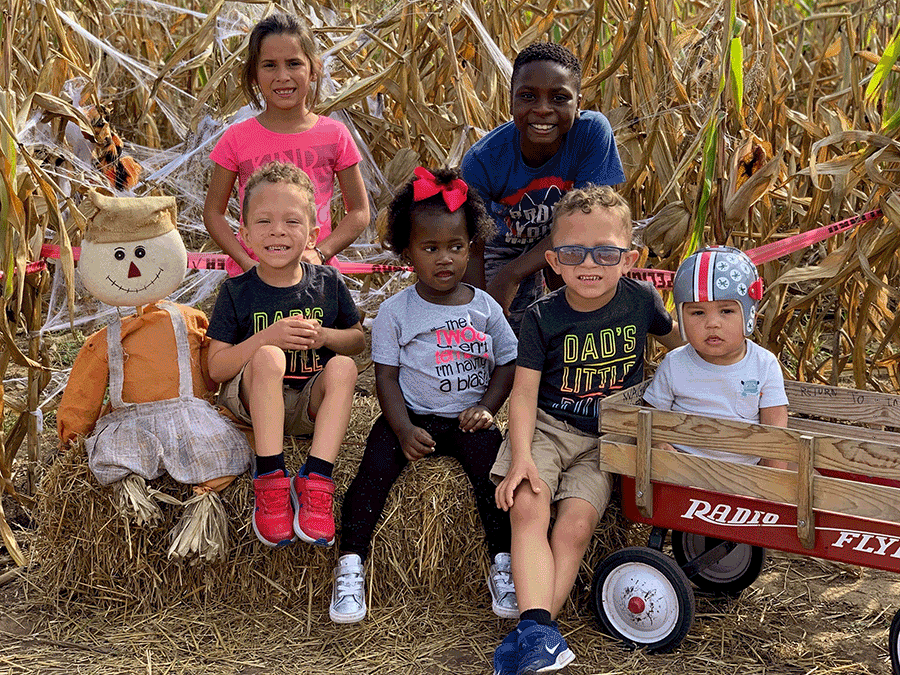 The six Keller children sitting in front of corn field on hay with scarecrow in fall. 