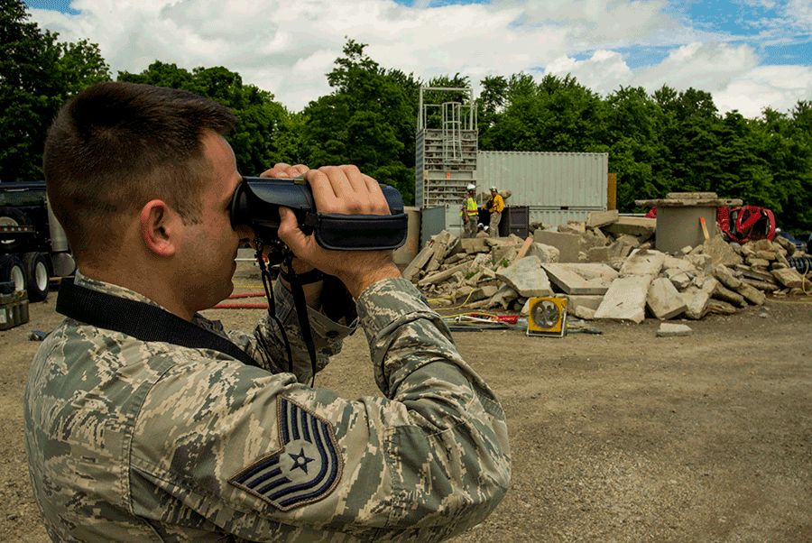 Airman looks through binoculars at disaster area with rubble in background. 