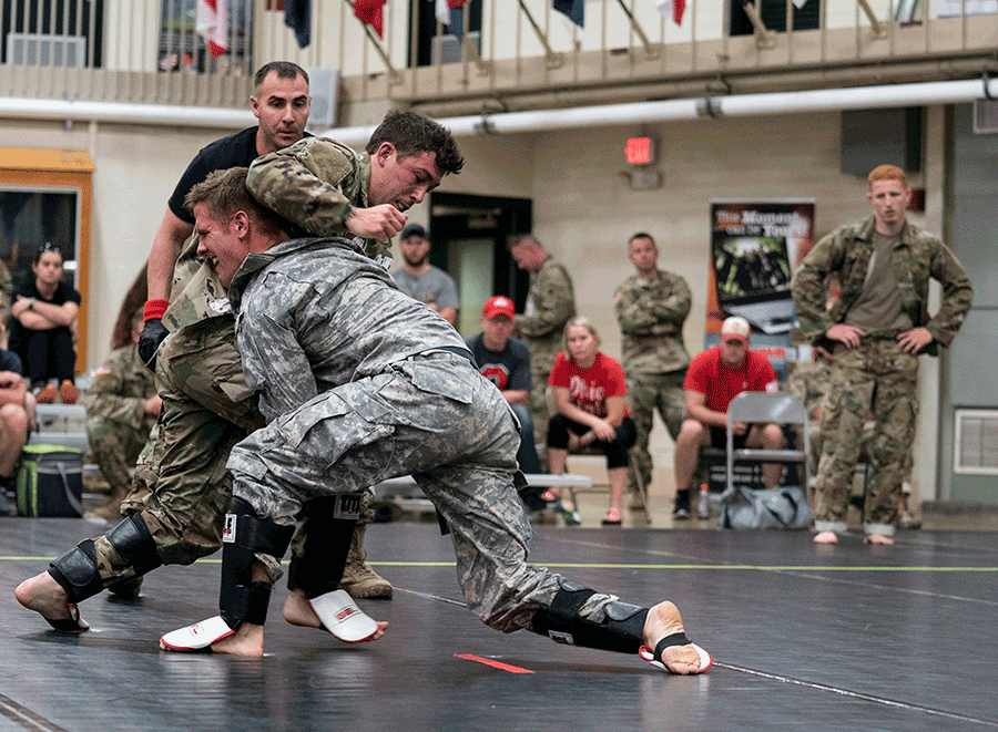 Ref oversees Soldiers fighting on mat while spectators watch from bleachers.