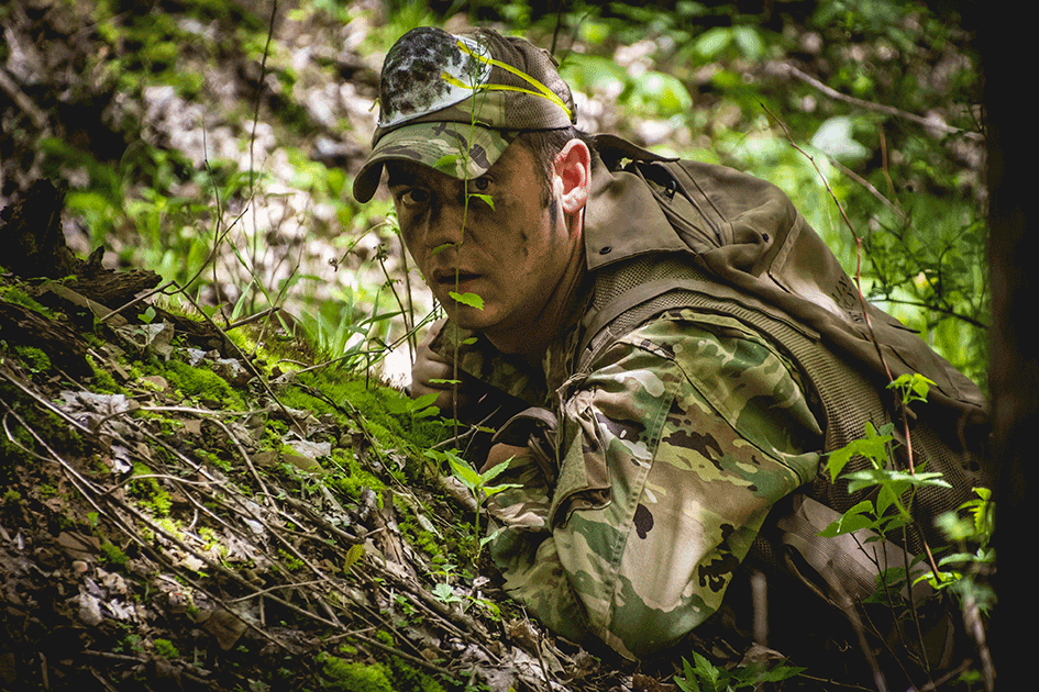Airman in full camo hides on ground in forest.