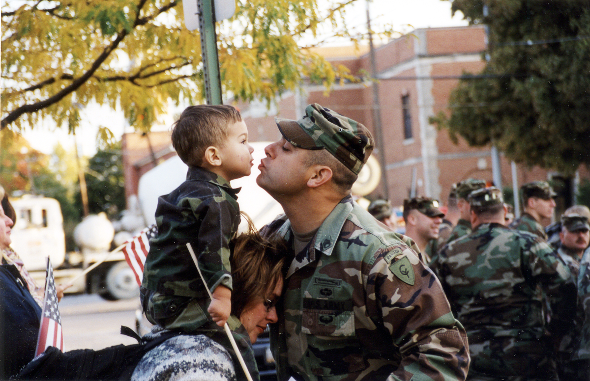 Soldier kisses son on top of wife's shoulders.