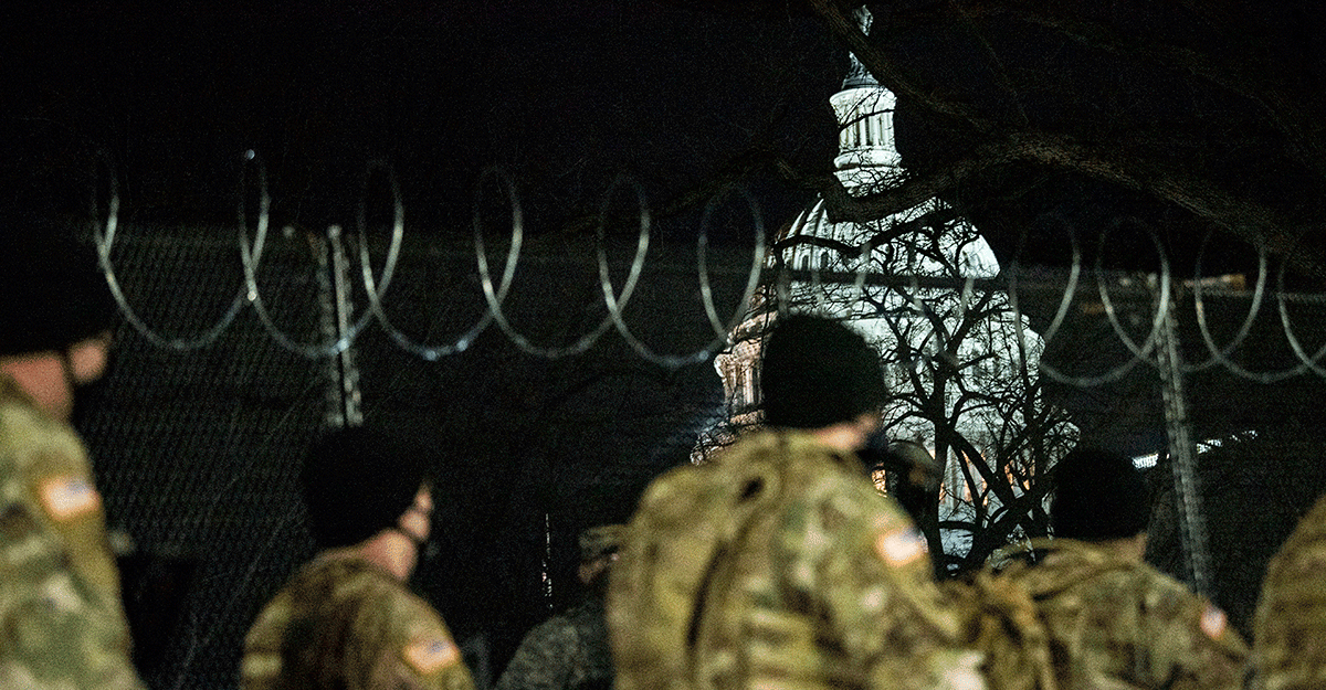 Members walk along barbed-wire fence at night with Capitol lit up.