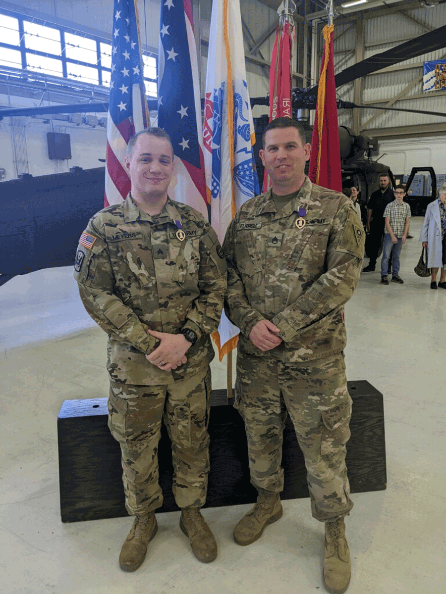 Two Soldiers stand in front of flags with their purple hearts pinned on.