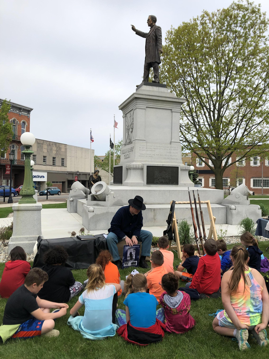 Mann in Civil War uniform talking with children at statue.
