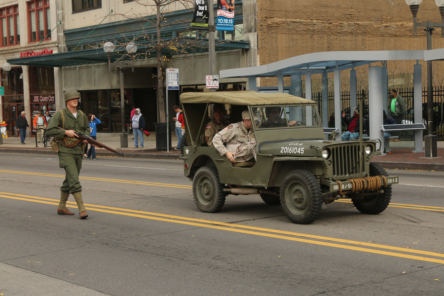 Mann marching in vintage uniform behind jeep in parade.