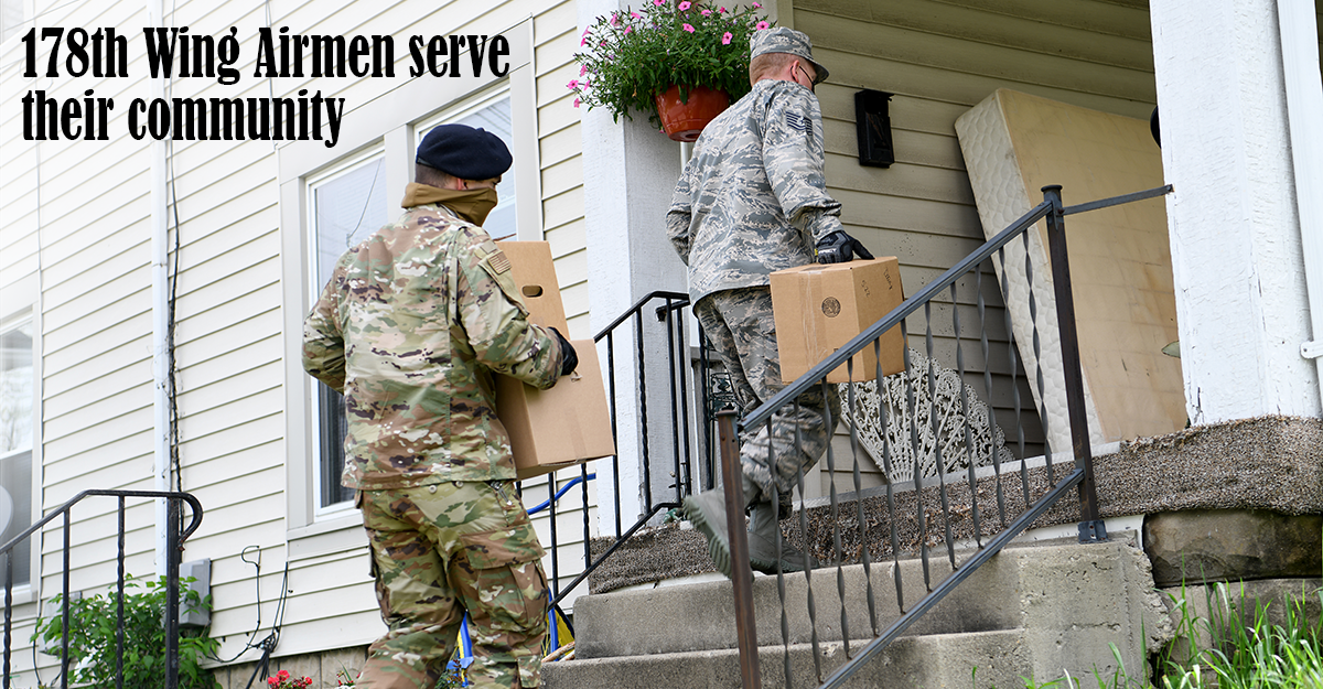 Two guard members carry boxes of supplies up steps to a beige house.